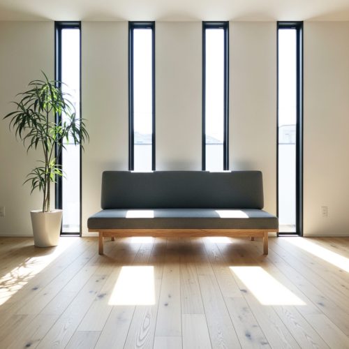 縦長のスリット窓から明るい自然光が差し込むリビング。無垢材のフローリングに置かれたグレーのソファと観葉植物が、心地よい空間を演出している。